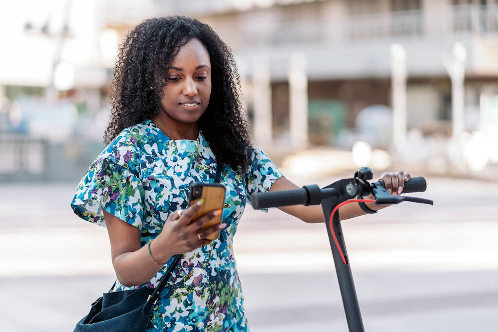 Heureuse femme noire avec scooter électrique marchant et utilisant son téléphone portable dans la rue. - Photo, image