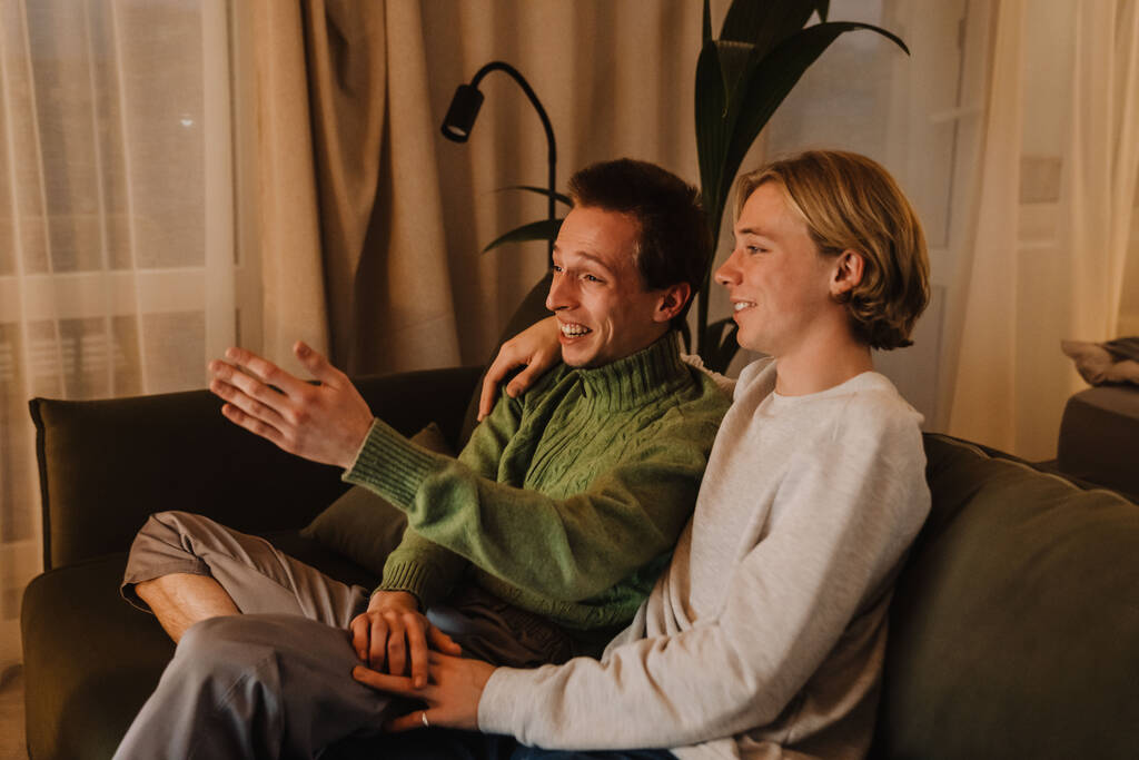 młody gej para śmiejąc się i odpoczywając na kanapie podczas oglądanie TV w domu - Zdjęcie, obraz