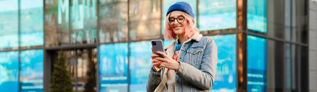 屋外の街の通りに立っている間、携帯電話を使用してメガネの若い女性 - 写真・画像