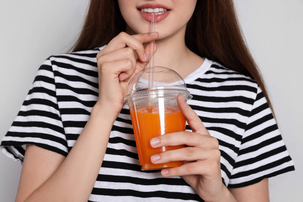 Νεαρή γυναίκα πόσιμο χυμό από πλαστικό κύπελλο σε ανοιχτό γκρι φόντο, closeup - Φωτογραφία, εικόνα