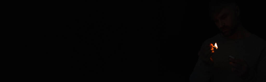 homem com caixa de fósforos olhando para chama de fósforo iluminado isolado em preto, banner - Foto, Imagem