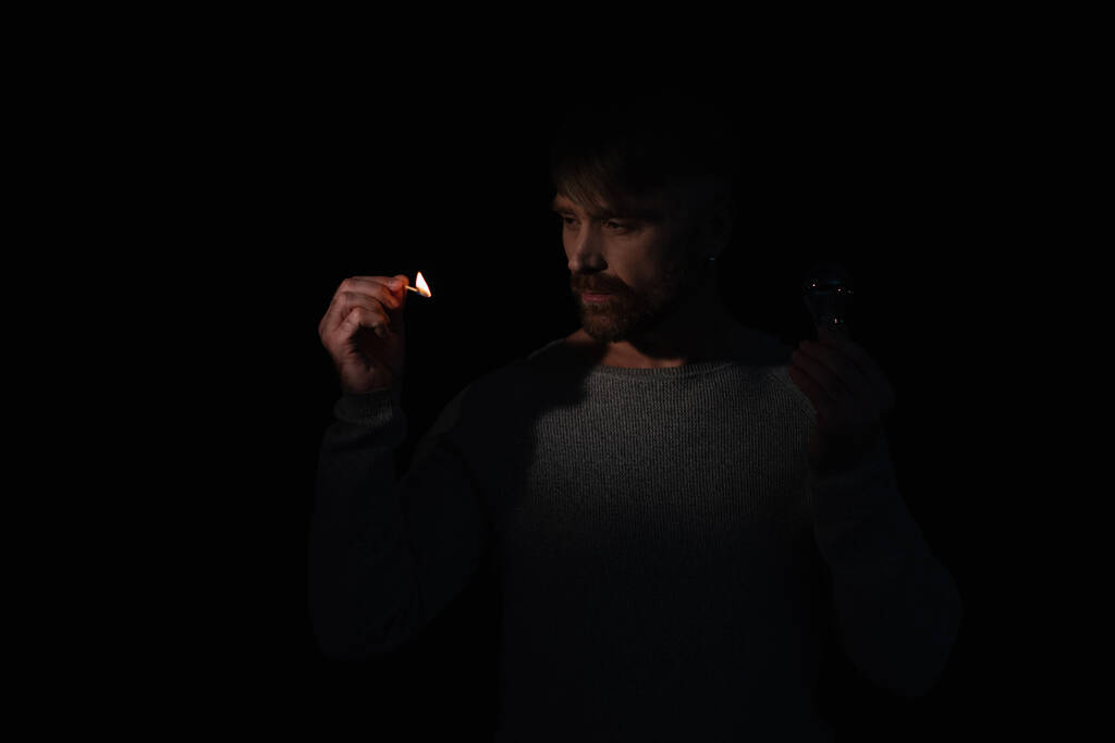 muž ve tmě drží elektrickou žárovku a dívá se na plamen zapálené zápalky izolované na černé - Fotografie, Obrázek