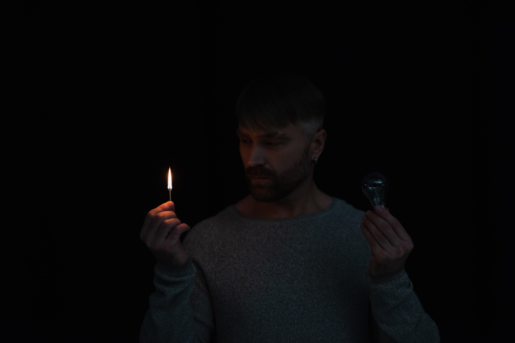 man looking at flame of lit match while holding electric bulb isolated on black - Photo, Image