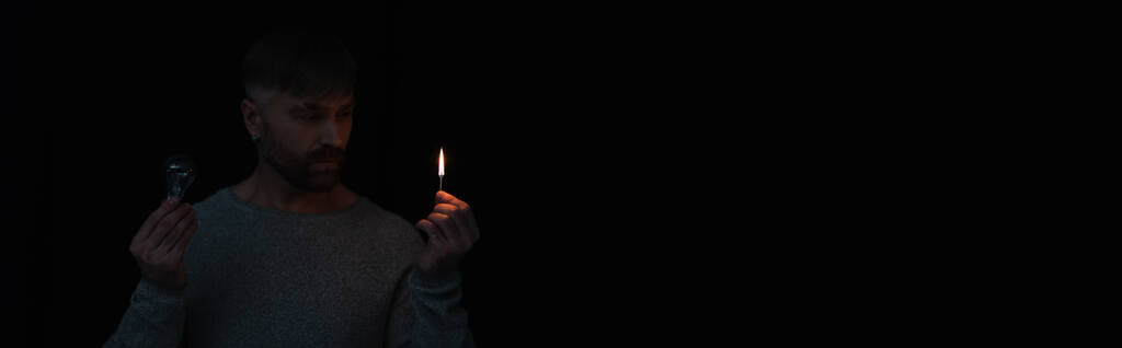 Mann während Stromabschaltung hält brennendes Streichholz und Glühbirne isoliert auf schwarz, Banner - Foto, Bild