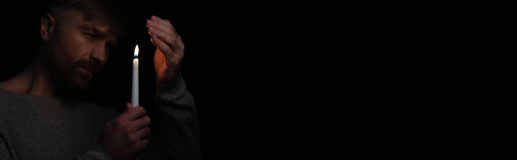 hombre de la mano cerca de la vela encendida durante el apagón eléctrico aislado en negro, pancarta - Foto, imagen