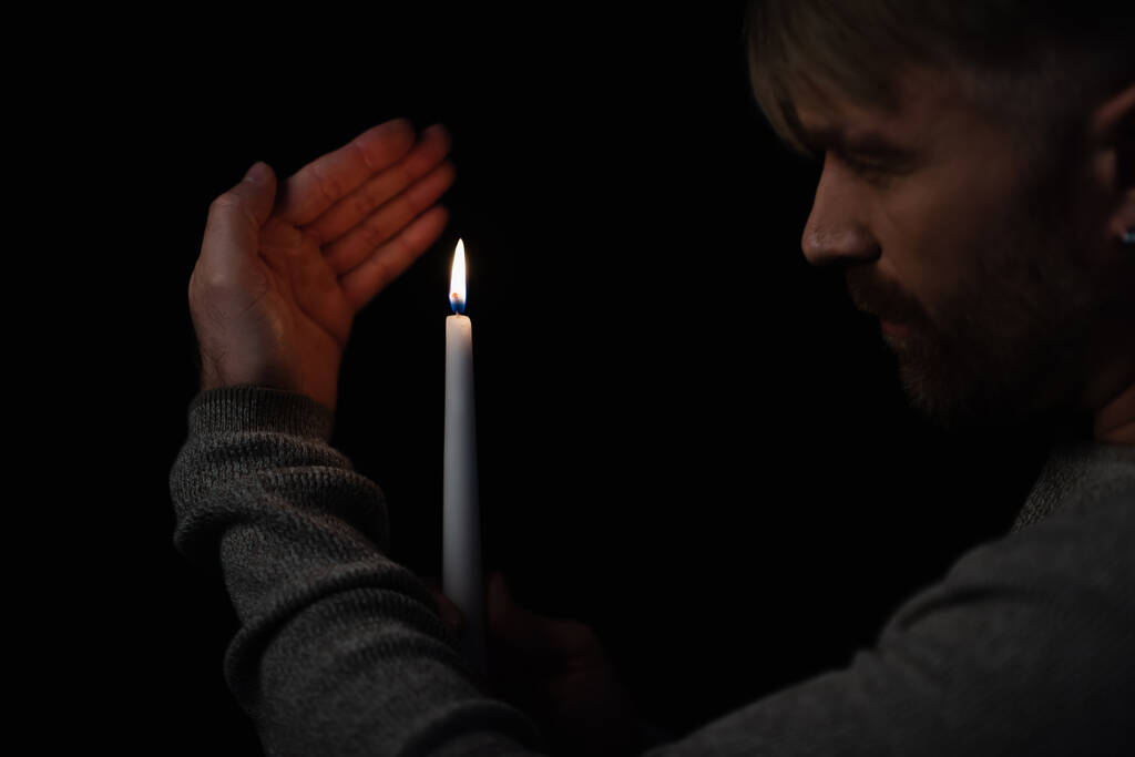 hombre mirando la llama de la vela encendida durante el corte de energía aislado en negro - Foto, Imagen