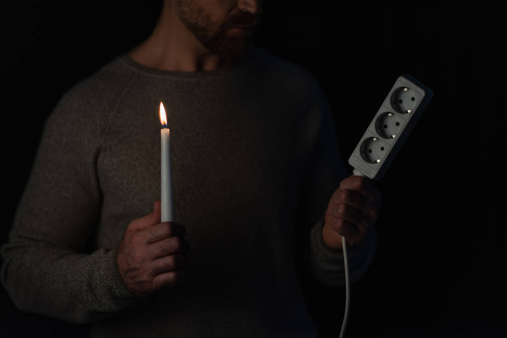 частичное представление человека, держащего удлинитель розетки и зажжённую свечу во время отключения электроэнергии, изолированного на черном - Фото, изображение