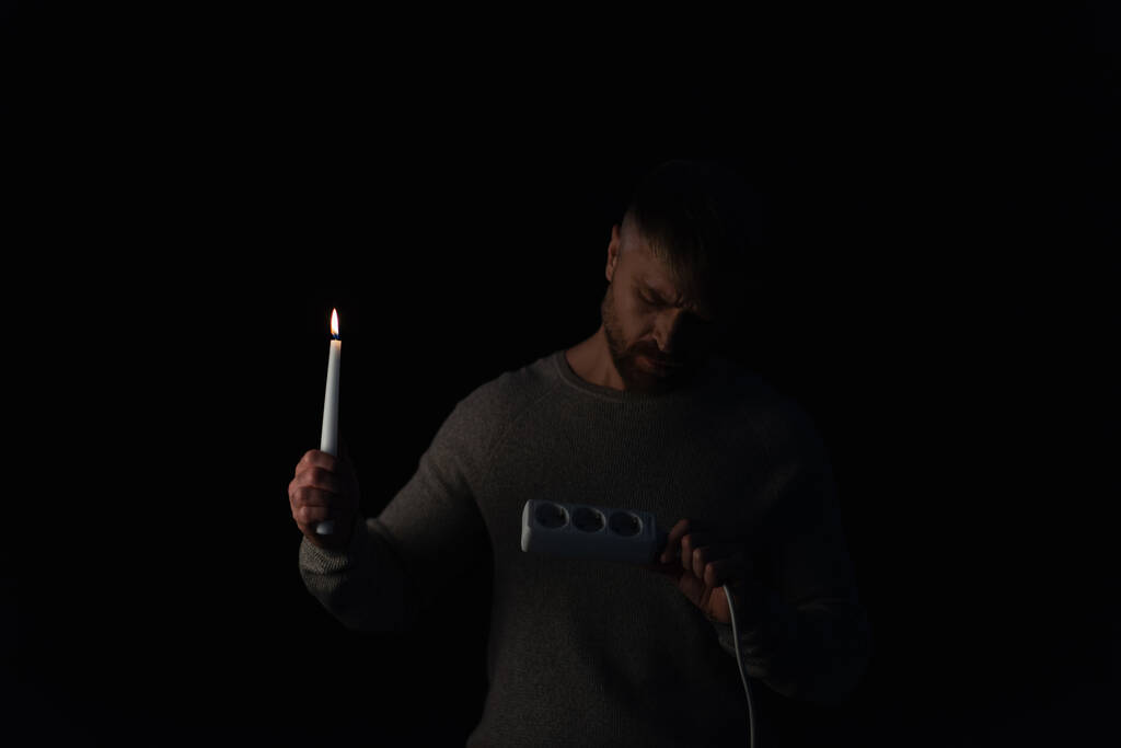 Mann mit brennender Kerze schaut bei Stromausfall auf Steckdosenverlängerer - Foto, Bild