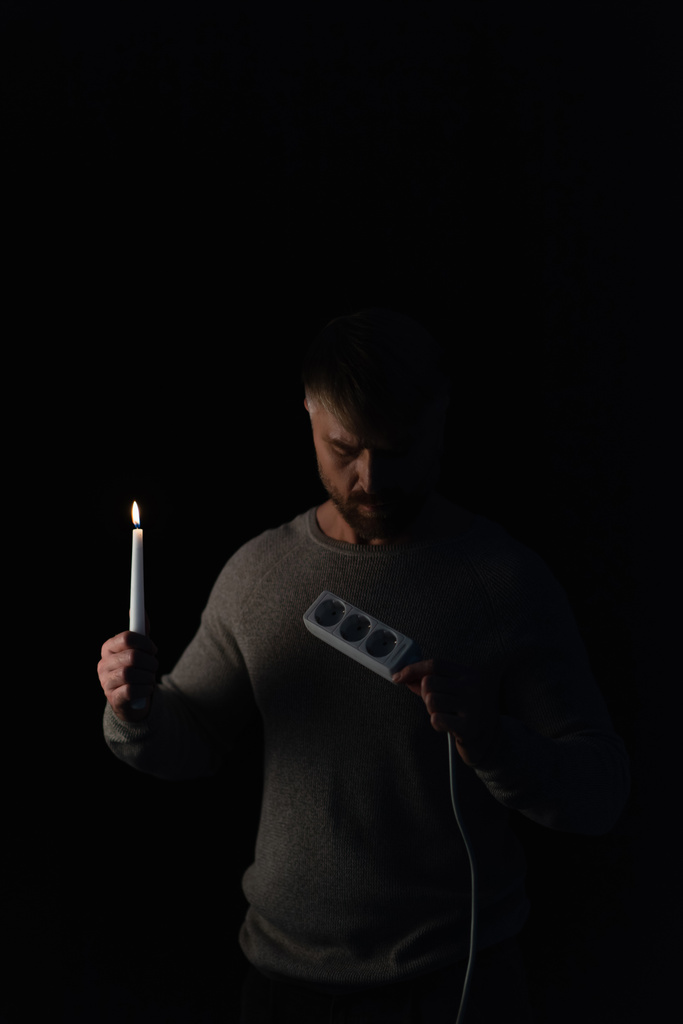 hombre adulto sosteniendo vela encendida en la oscuridad y mirando extensor de enchufe aislado en negro - Foto, imagen