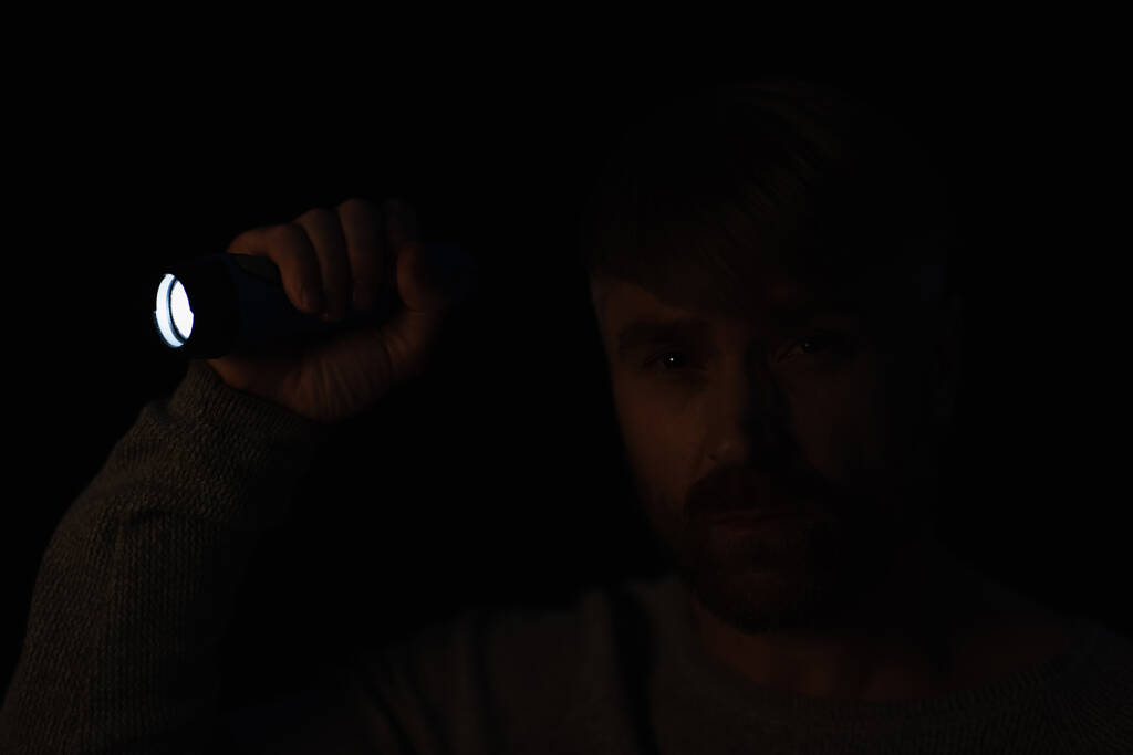 homem na escuridão olhando para a câmera enquanto segurando lanterna brilhante isolado no preto - Foto, Imagem
