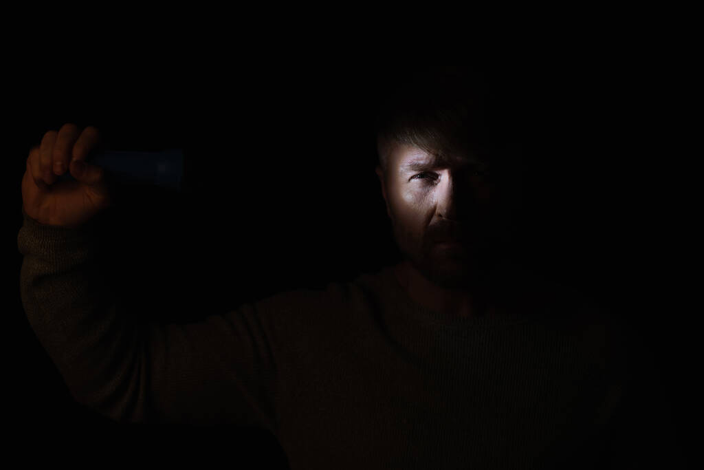 homme dans l'obscurité éclairage sur le visage avec une lampe de poche et en regardant la caméra isolée sur noir - Photo, image