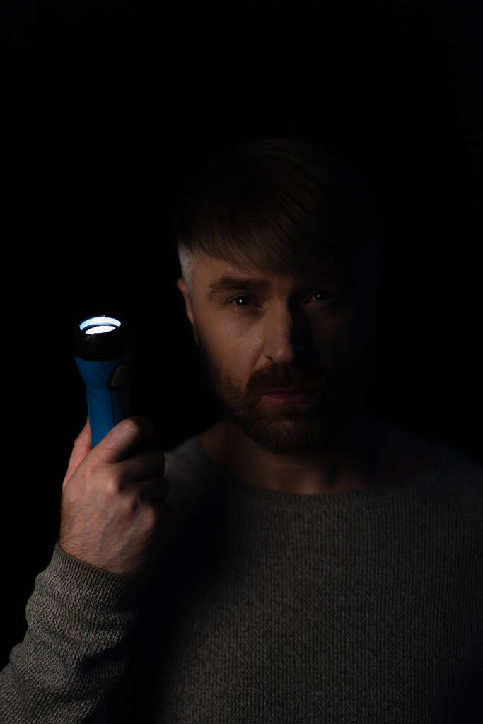 hombre con linterna brillante mirando a la cámara durante el apagado de electricidad aislado en negro - Foto, Imagen