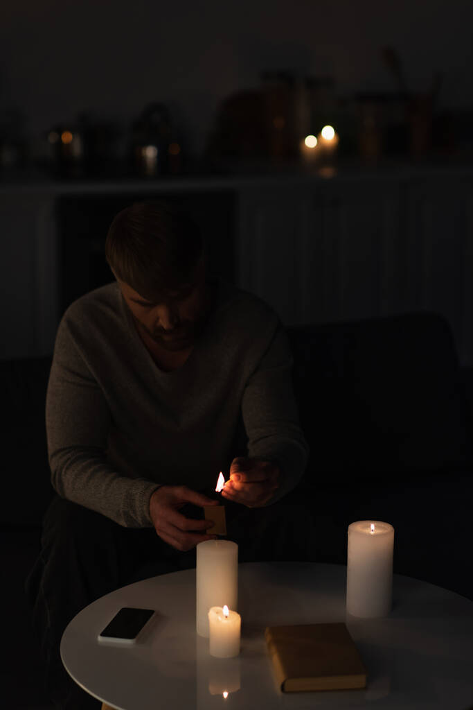 человек, сидящий в темноте и зажигая свечи возле книги и мобильного телефона с пустым экраном - Фото, изображение