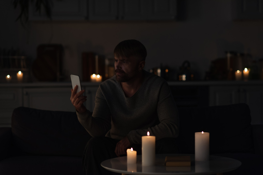 mies katselee matkapuhelinta istuessaan pimeässä keittiössä lähellä polttavia kynttilöitä  - Valokuva, kuva