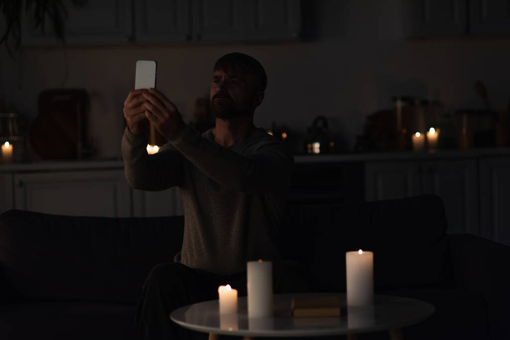 mies istuu lähellä polttava kynttilöitä pimeässä keittiössä ja kiinni mobiili yhteys älypuhelimeen - Valokuva, kuva
