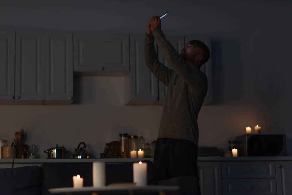 man houdt mobiele telefoon in opgeheven handen, terwijl het vangen van mobiele signaal in de donkere keuken in de buurt van brandende kaarsen - Foto, afbeelding