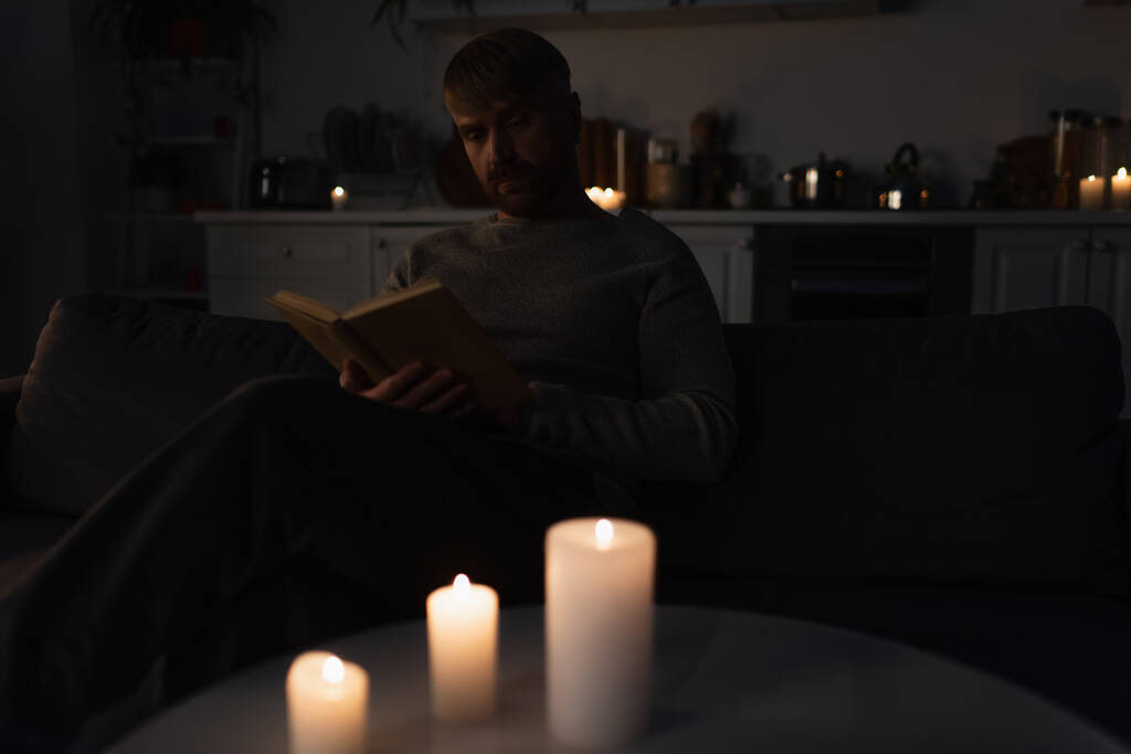 hombre sentado en la cocina durante el apagón de energía y leer libro cerca de velas encendidas  - Foto, imagen