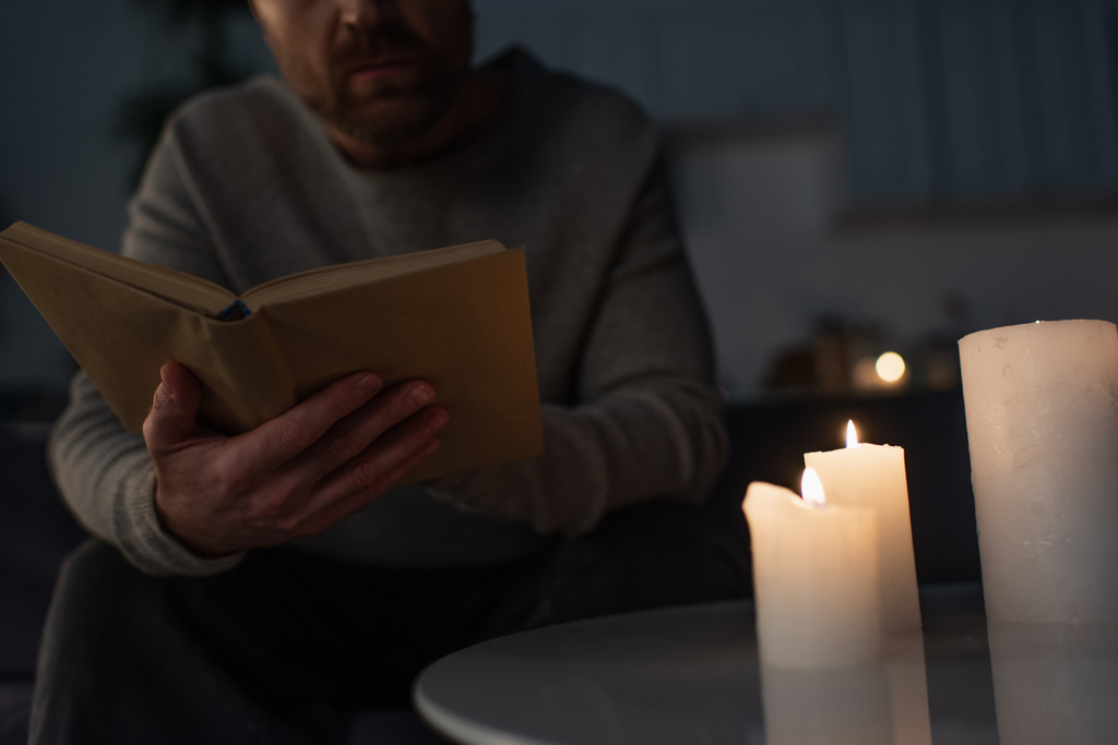 oříznutý pohled na muže čtení knihy u svíček hořící v tmavé kuchyni během výpadku proudu - Fotografie, Obrázek