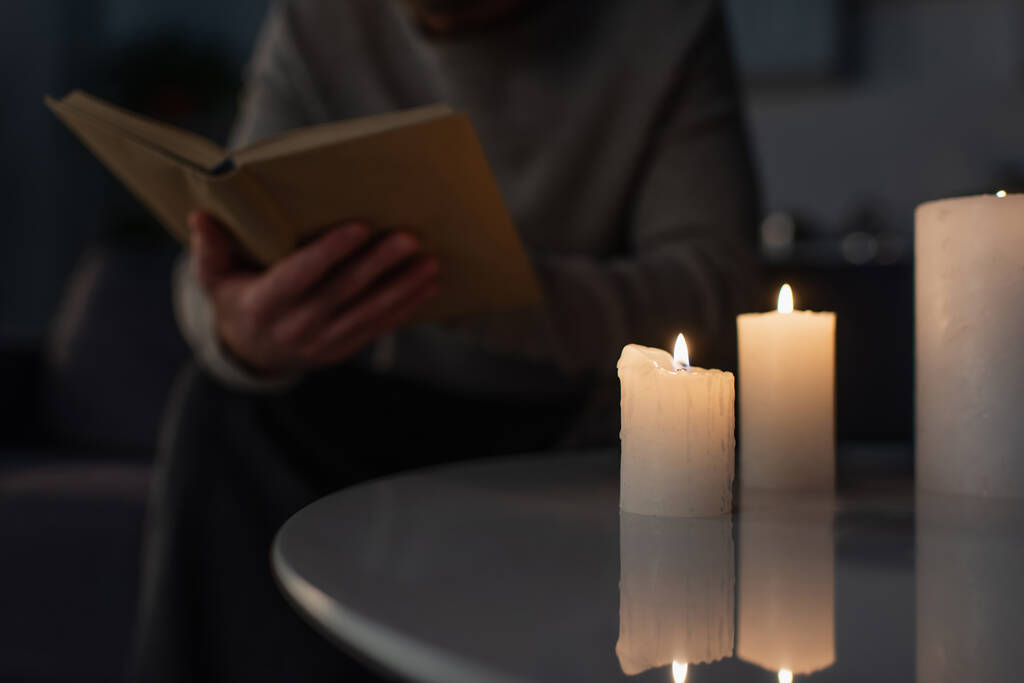 vista parcial del hombre borroso leyendo libro en la oscuridad cerca de encender velas en la mesa - Foto, Imagen