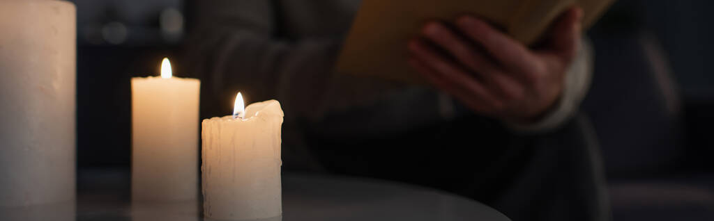 επιλεκτική εστίαση της καύσης κεριών κοντά περικοπεί άνθρωπος ανάγνωση βιβλίο στο σκοτάδι σε θολή φόντο, πανό - Φωτογραφία, εικόνα