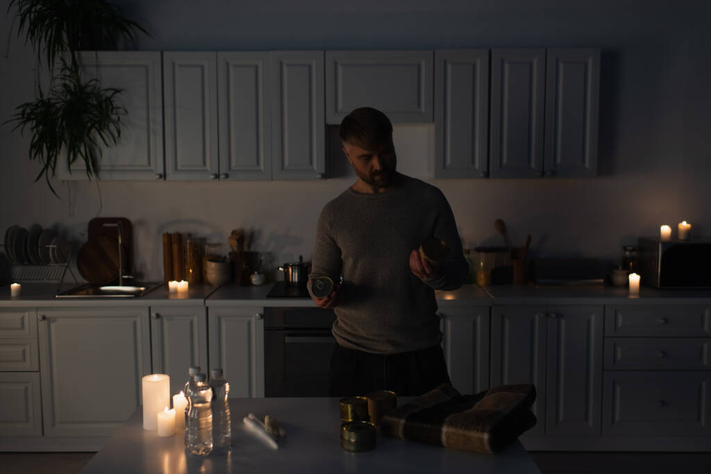 Mann hält Konserven neben Tisch mit Mineralwasser und Kerzen  - Foto, Bild