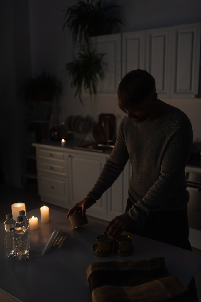 homme tenant de la nourriture en conserve près de l'eau embouteillée et des bougies dans la cuisine pendant la panne d'électricité - Photo, image