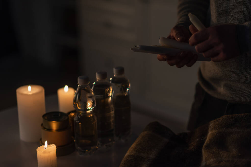 osittainen näkymä miehestä, jolla on kynttilöitä lähellä pullotettua vettä säilykkeiden ja lämpimän peiton kanssa pöydällä - Valokuva, kuva