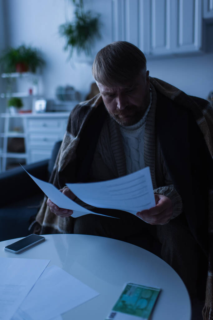 homem sentado sob cobertor quente e olhando para faturas perto de dinheiro e smartphone durante o desligamento de energia - Foto, Imagem