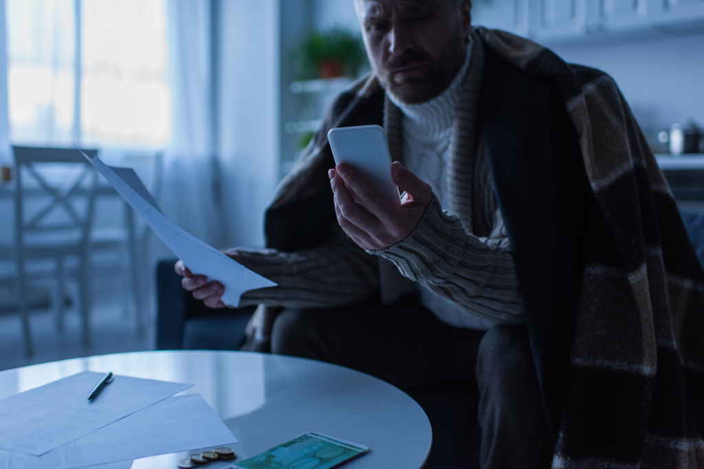 zamrożony człowiek z fakturami patrząc na smartfona blisko pieniędzy na stole w zmierzchu - Zdjęcie, obraz