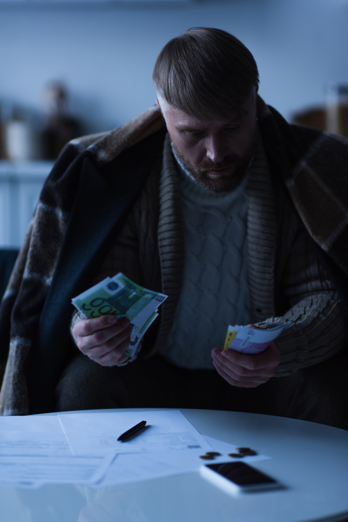 человек под теплым одеялом подсчитывает банкноты евро возле платежных счетов во время отключения электроэнергии - Фото, изображение