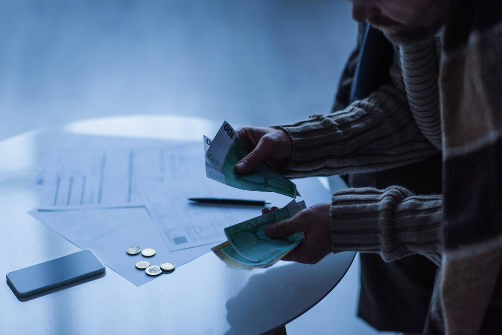 частичное представление человека, подсчитывающего деньги возле смартфона и платежные счета в сумерках - Фото, изображение