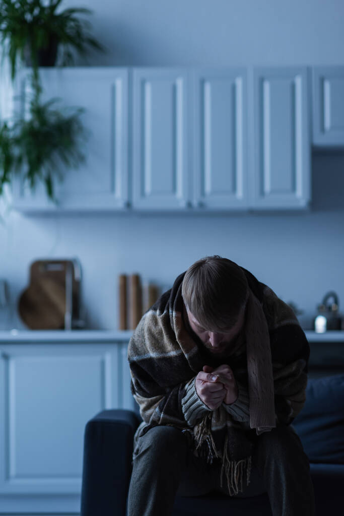 Mann in Decke gehüllt wärmende Hände, während er während des Stromausfalls in der Küche sitzt - Foto, Bild