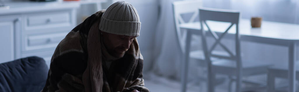 Замороженный человек сидит в теплом одеяле и вязаной шляпе дома в сумерках, баннер - Фото, изображение