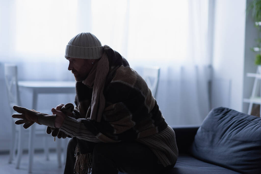 side view of frozen man putting on gloves while sitting on couch in hat and warm blanket - Photo, Image