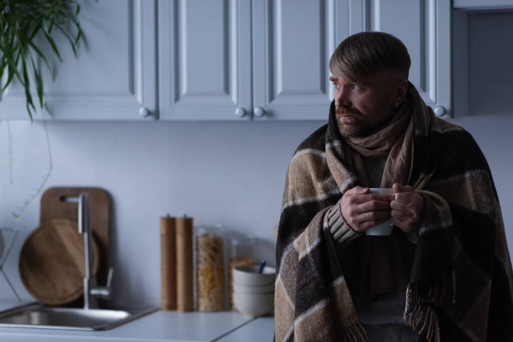Sıcak battaniyeli donmuş bir adam elinde bir bardak sıcak içecekle mutfağa bakıyor. - Fotoğraf, Görsel
