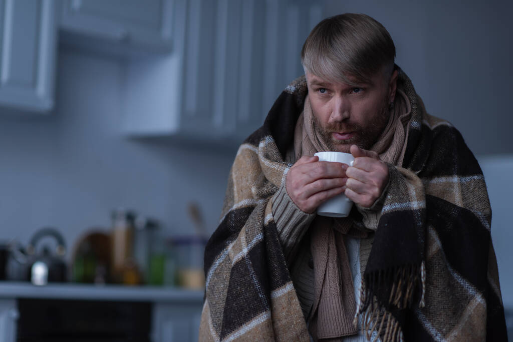 Mann steht bei Stromausfall zu Hause unter warmer Decke und bläst heißen Tee - Foto, Bild