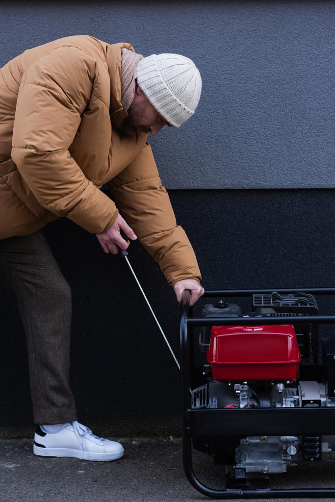 Seitenansicht eines Mannes in Daunenjacke und Strickmütze, der im Freien einen Stromgenerator startet - Foto, Bild