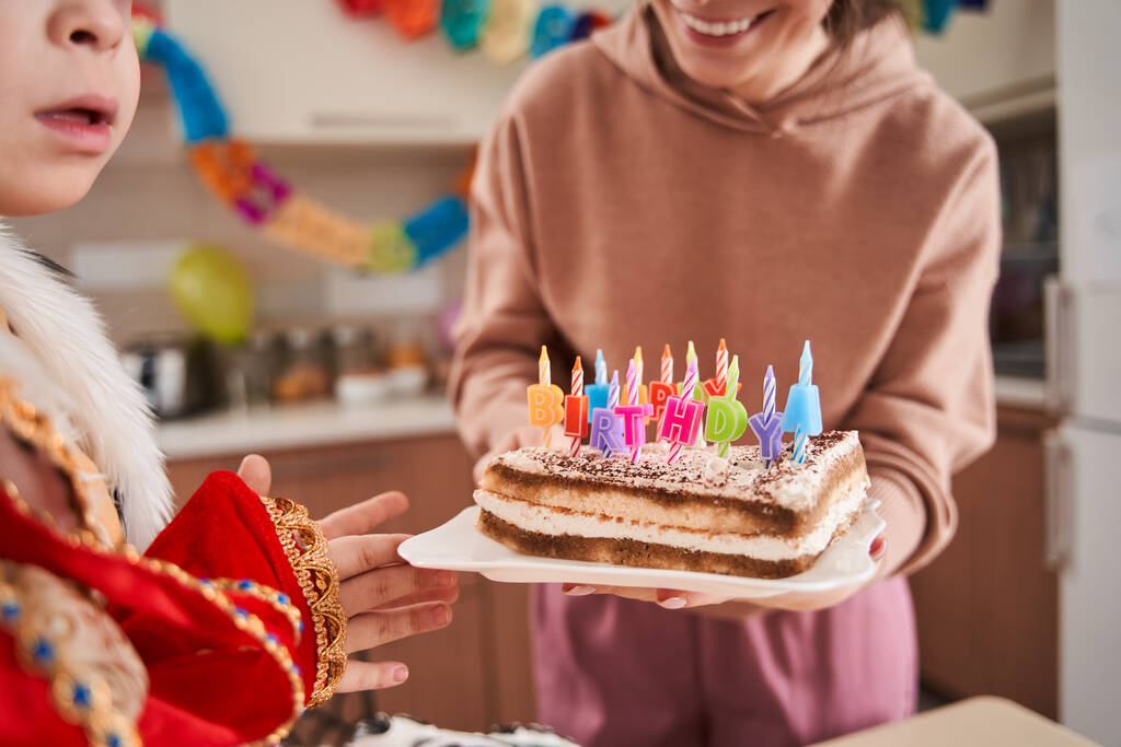 Vista ritagliata della donna caucasica con torta di compleanno per mano in piedi vicino al suo bambino con sindrome di Down in cucina - Foto, immagini