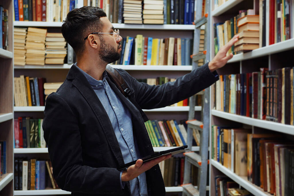 Homem com barba e óculos vestindo camisa e jaqueta andando por estantes e escolhendo livros, tablet com lista de literatura em sua mão, mochila no ombro. Estudante universitário em biblioteca - Foto, Imagem