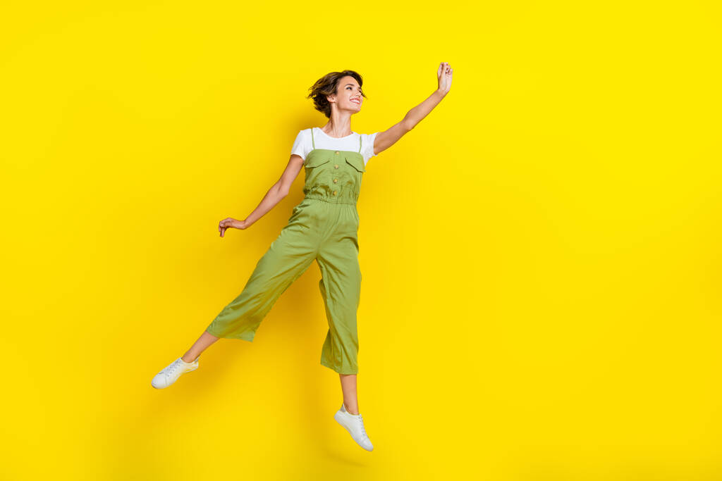 Foto de cuerpo completo de joven linda mujer traje de verano llevar overoles saltar aire mantenga los dedos interesados espacio vacío aislado en el fondo de color amarillo. - Foto, imagen