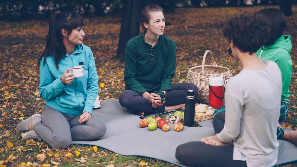 Piękny najmłodszych są relaks po treningu na zewnątrz pikniku na matach jedzenia przekąski i picia herbaty. Dziewczyny są rozmawiając i śmiejąc się korzystających jesień natura. - Zdjęcie, obraz
