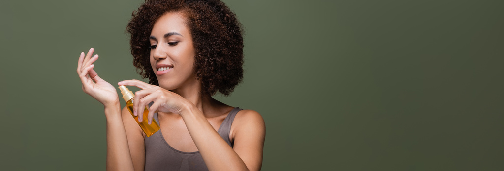 Vidám afro-amerikai nő alkalmazó kozmetikai olaj a kezét elszigetelt zöld, banner  - Fotó, kép