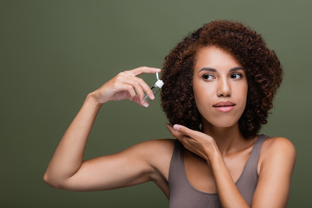 Afroamerikanerin in Top hält Kosmetiköl in der Nähe lockiges Haar isoliert auf grün  - Foto, Bild