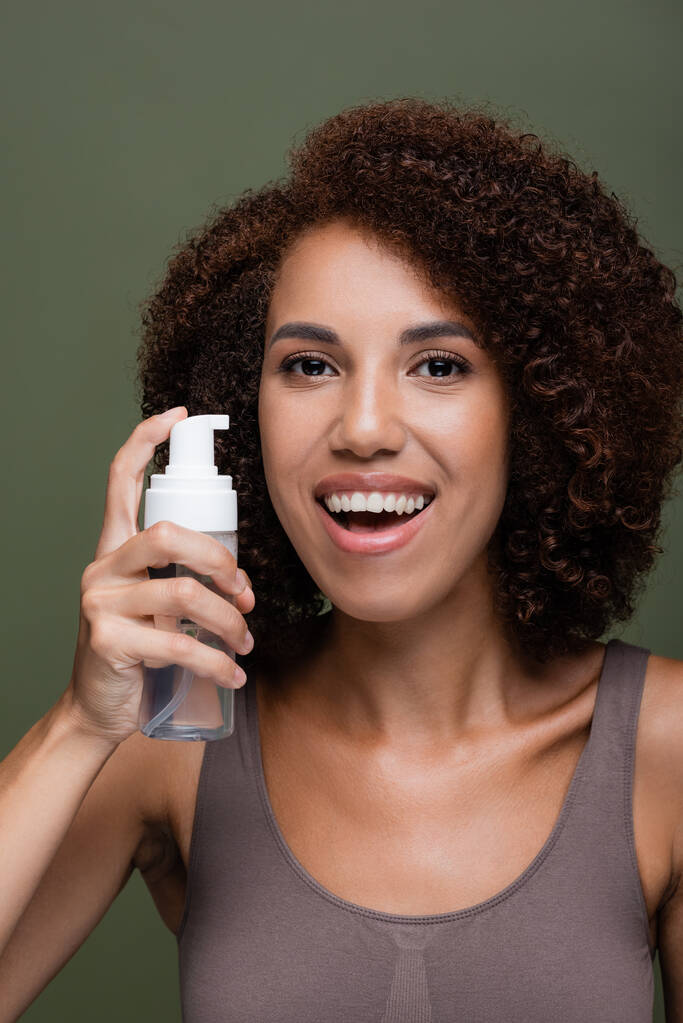 Porträt einer lächelnden Afroamerikanerin, die Kosmetikschaum in der Hand hält und isoliert auf Grün in die Kamera blickt  - Foto, Bild