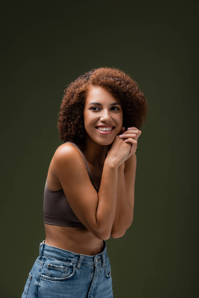 Joyeux et frisé modèle afro-américain en haut et jeans regardant loin isolé sur vert foncé  - Photo, image