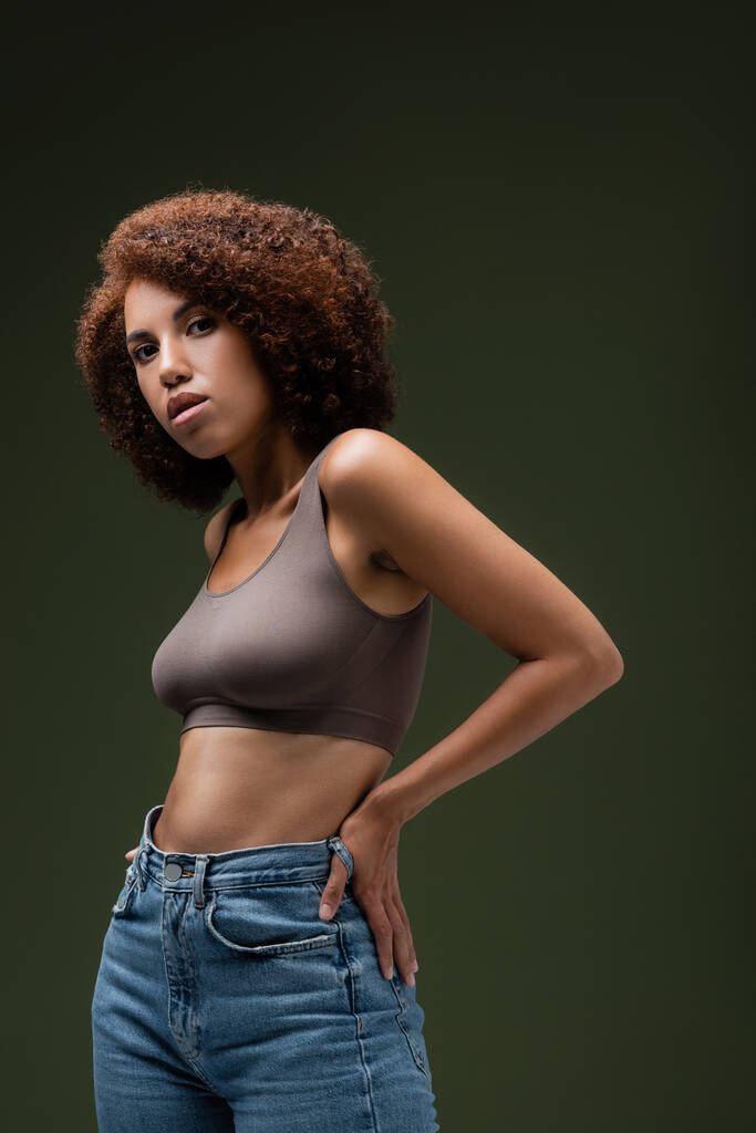 Curly african american woman in top and jeans holding hand on hip and looking at camera isolated on dark green  - Photo, Image