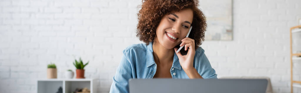 Femme afro-américaine positive parlant sur un téléphone portable près d'un ordinateur portable flou à la maison, bannière  - Photo, image