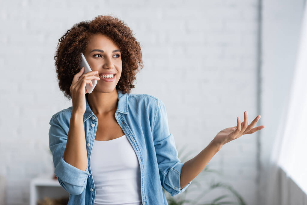 boldog afro-amerikai nő beszél okostelefonon és gesztikulál otthon  - Fotó, kép
