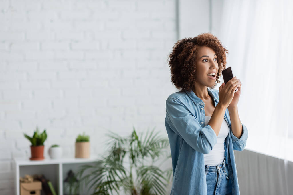 mujer afroamericana emocionada sosteniendo teléfono inteligente con pantalla en blanco y mirando hacia otro lado - Foto, imagen