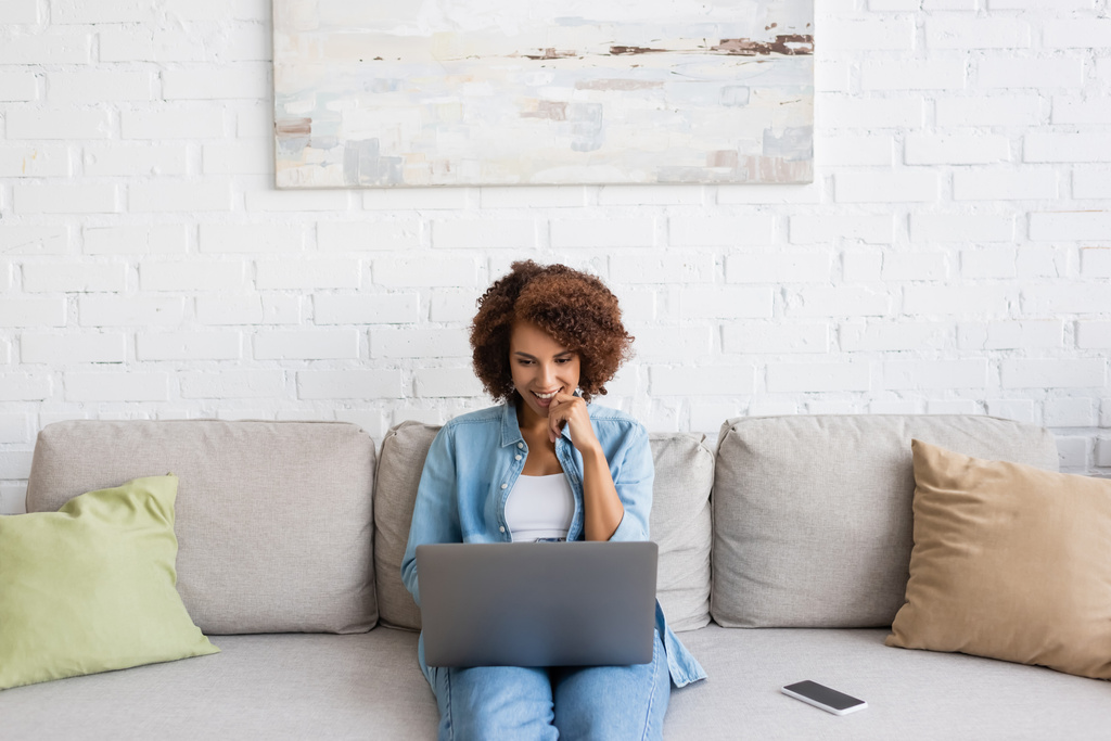 felice donna afroamericana utilizzando il computer portatile mentre seduto sul divano e lavorare da casa  - Foto, immagini